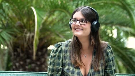 Beautiful-young-woman-listening-to-music-in-the-park-against-the-backdrop-of-a-palm-tree-and-waving-back-with-a-smile-to-someone