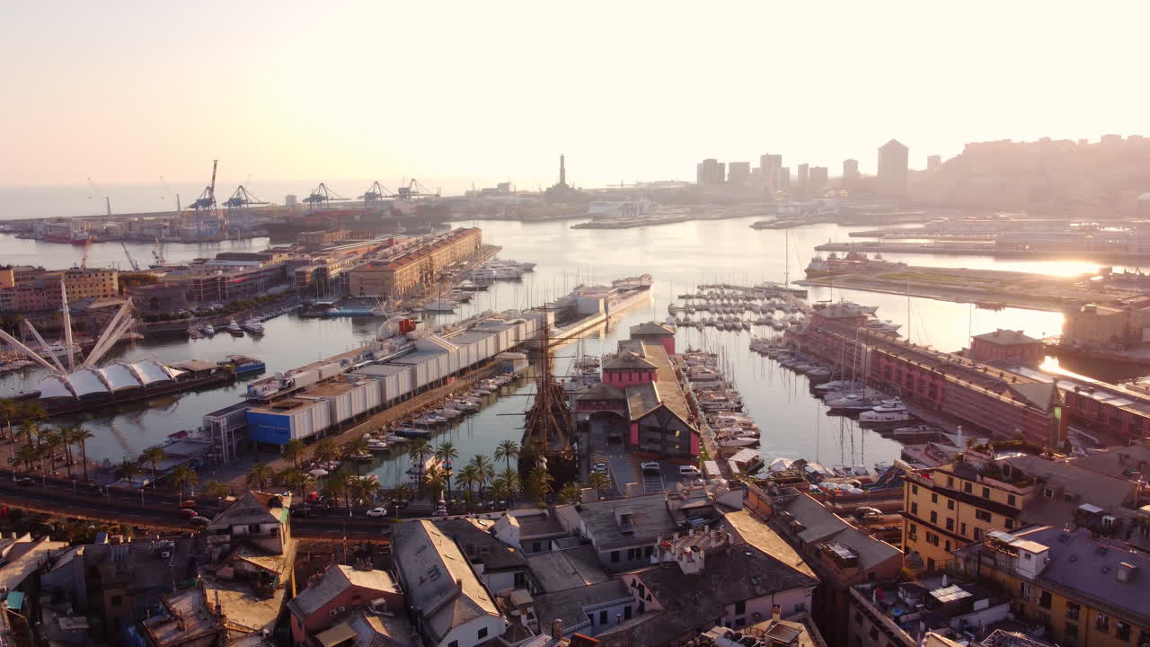 Drone panning shot showing Genoa port with boats, cranes, and waterfront structures, including part of the historical center at sunset
