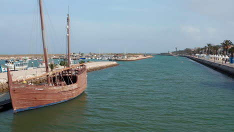 Lagos-harbor-canal-with-wooden-boat-Portuguese-caravela-anchored-on-dock.-Aerial-drone-flying-above-forward-above-water,-Portugal,-day