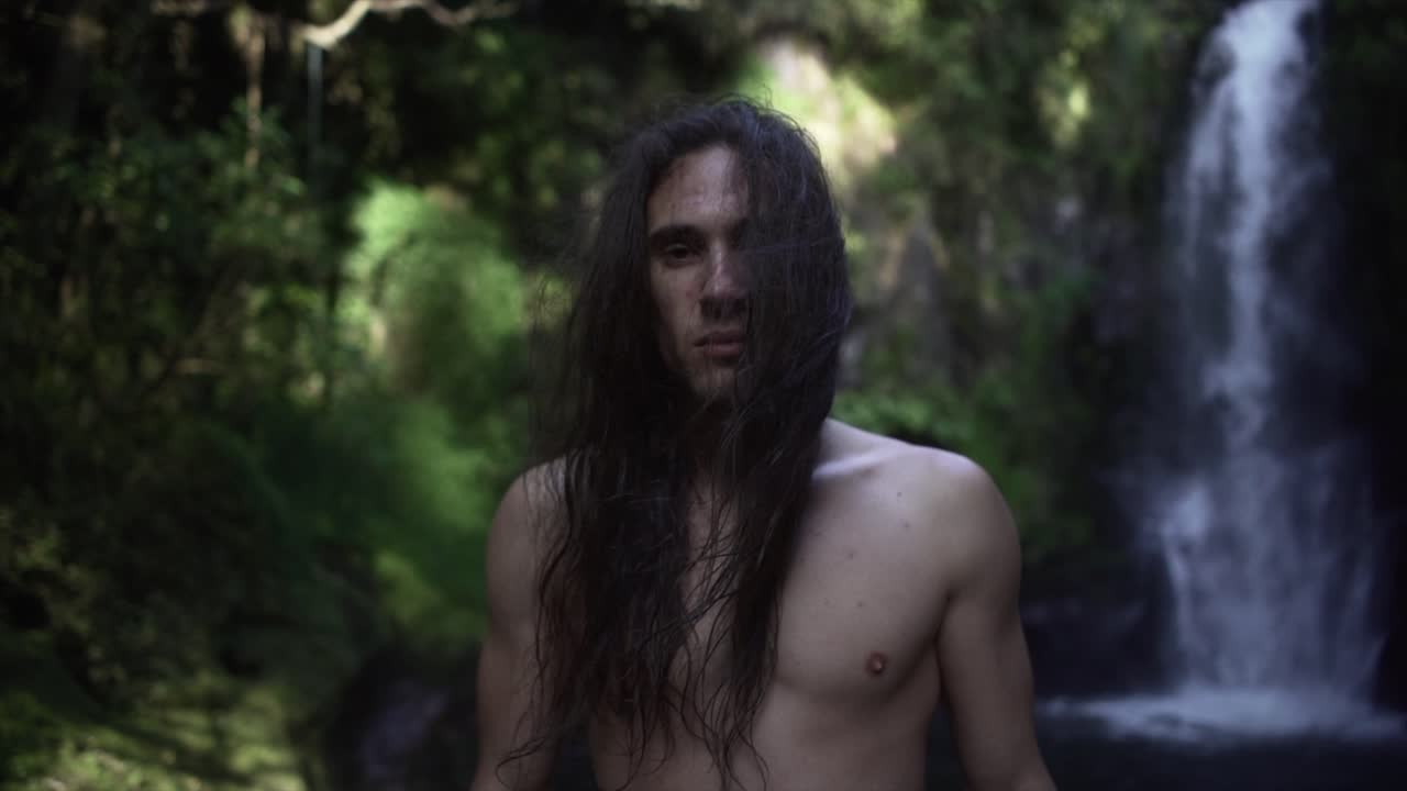 Man with long hair in front of a waterfall