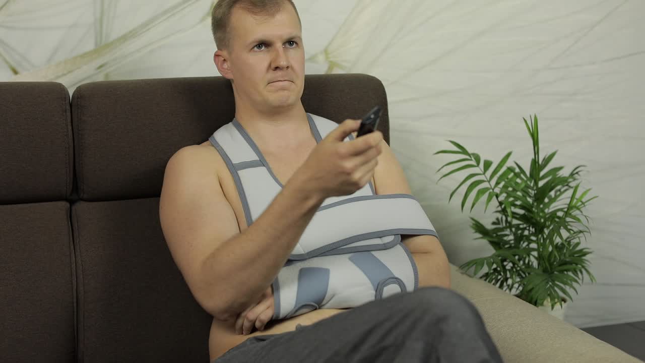 hombre doloroso con un brazo roto usando un soporte para el brazo sentado en un sofá viendo la televisión