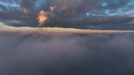 Toma-Aérea-De-Carro-Inverso-Sobre-Las-Nubes-Durante-La-Puesta-De-Sol-Con-Reflejos-Cálidos-Que-Contrastan-Con-El-Cielo-Azul-Y-Las-Nubes-Grises