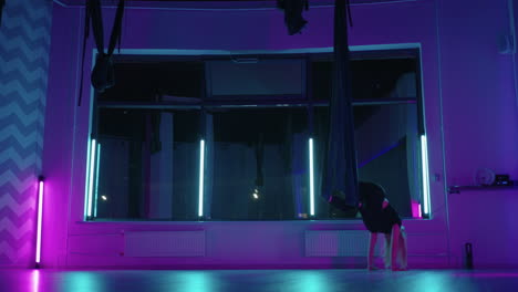 a woman does yoga and stretching on a hanging hammock in neon light. young pretty slim body fitness girl practicing fly yoga in the gym.