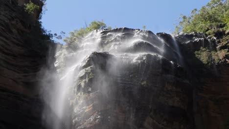 Vista-De-ángulo-Bajo-De-Las-Cataratas-De-Goingworth-En-Las-Montañas-Azules,-Australia-Con-El-Viento-Que-Sopla-El-Agua-Que-Baja-Y-El-Cielo-Azul