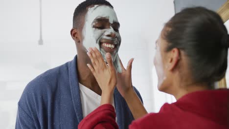 video de una feliz pareja diversa divirtiéndose aplicando máscaras faciales de limpieza y riendo en el baño