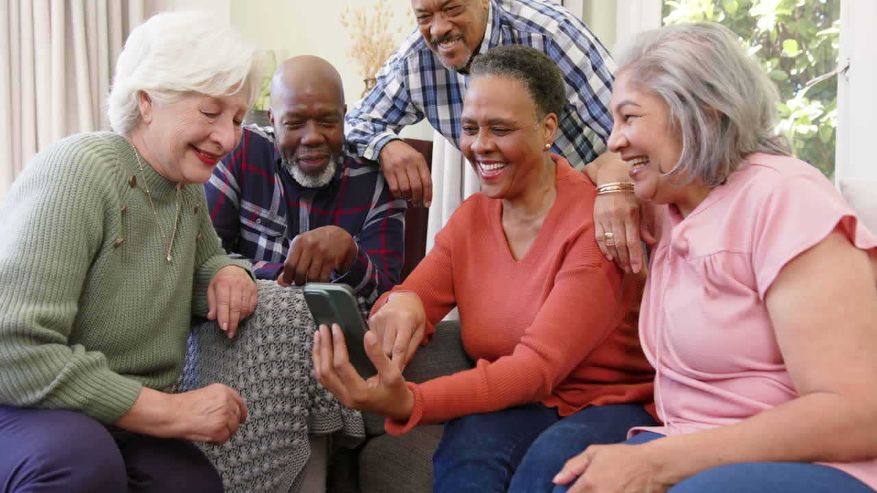 Amigos idosos felizes e diversos, homens e mulheres, usando smartphone juntos em uma sala ensolarada, em câmera lenta.