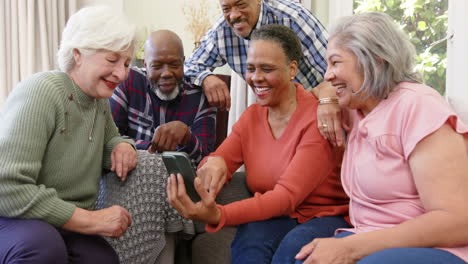 Happy-diverse-senior-male-and-female-friends-using-smartphone-together-in-sunny-room,-slow-motion