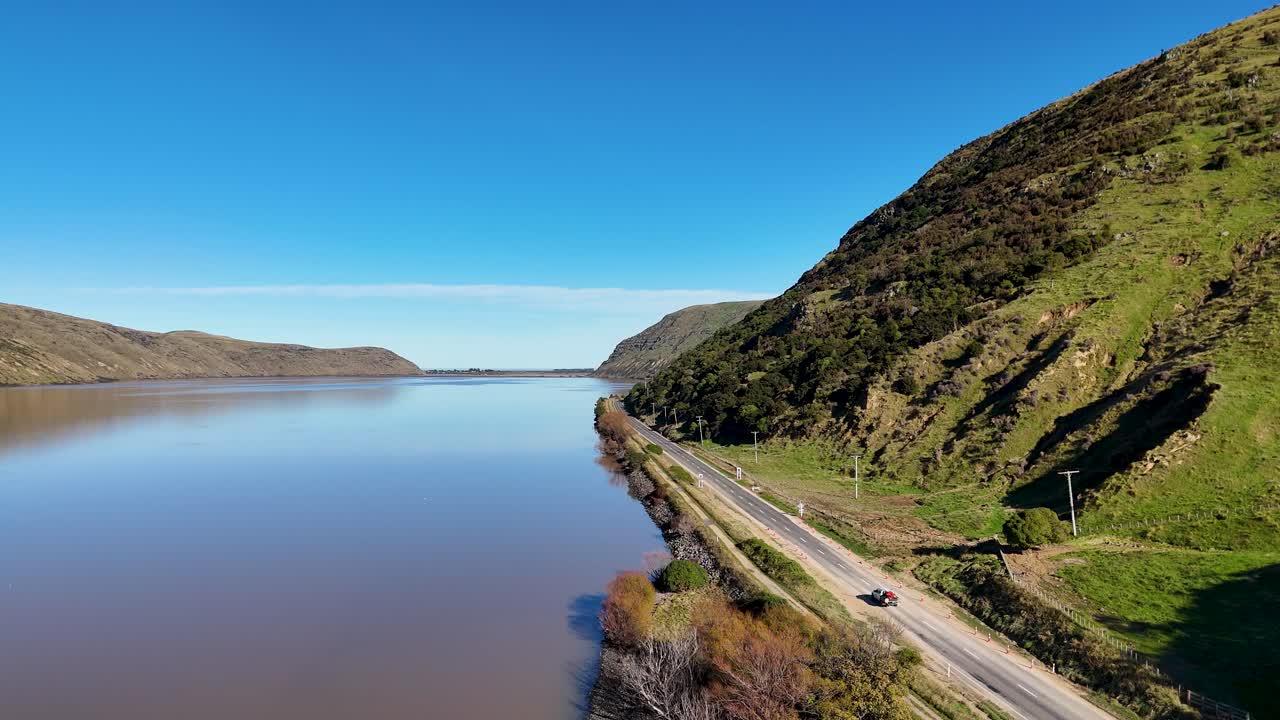 Aerial view of Akaroa's tranquil lake and winding road, captured in bright daylight with lush green hills