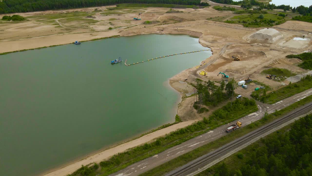 Video aereo di un drone su una grande cava di sabbia a cielo aperto, dove si trova un grande lago artificiale scavato di colore blu e un camion minerario sta guidando su una strada sabbiosa di ghiaia trasportando materiali. Attrezzature minerarie visibili
