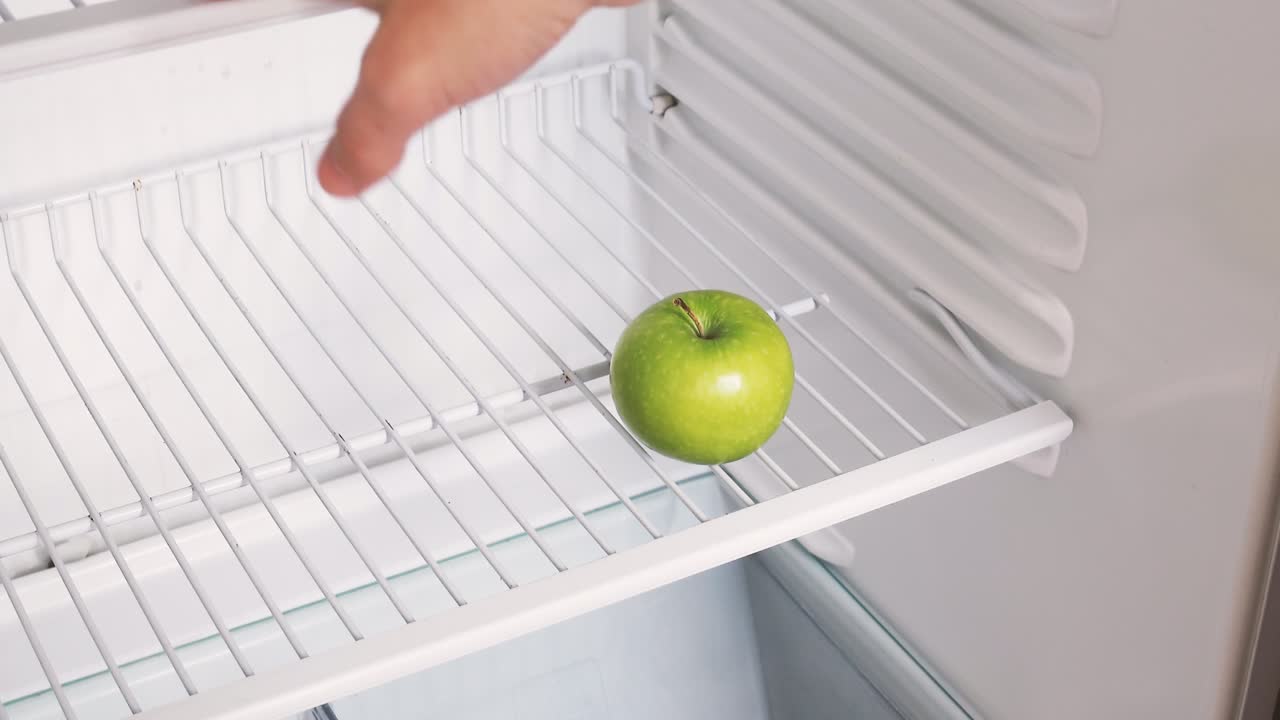 la mano de un hombre abre un refrigerador vacío y toma una manzana verde. video en bruto conceptual 4k. idea de crisis económica, escasez de alimentos, pobreza.