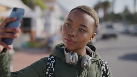 retrato de una estudiante afroamericana usando un teléfono inteligente posando tomando una foto selfie disfrutando de la actividad urbana en las vacaciones de verano