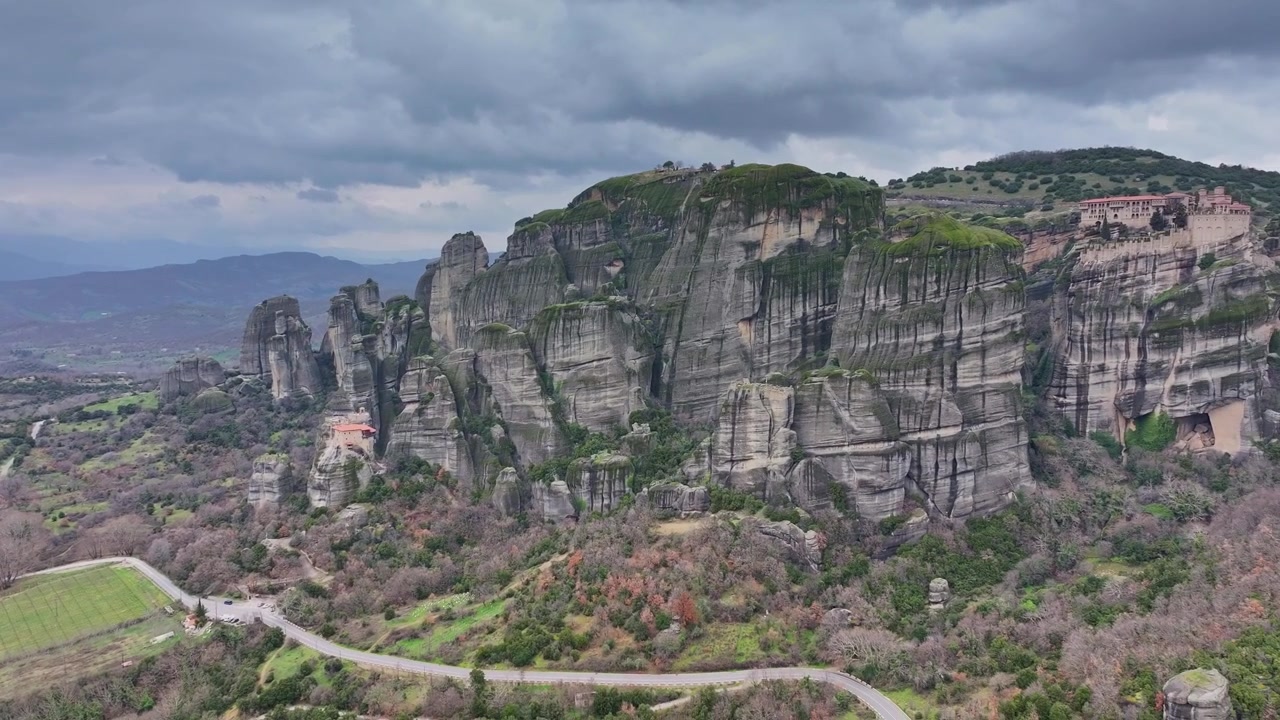 Tomada aérea de drone em panorâmica para a direita de um pilar de rocha para revelar os mosteiros de Roussanou e Varlaam empoleirados em penhascos maciços com estrada sinuosa e o vale da Tessália sob céu nublado em Meteora, Grécia.