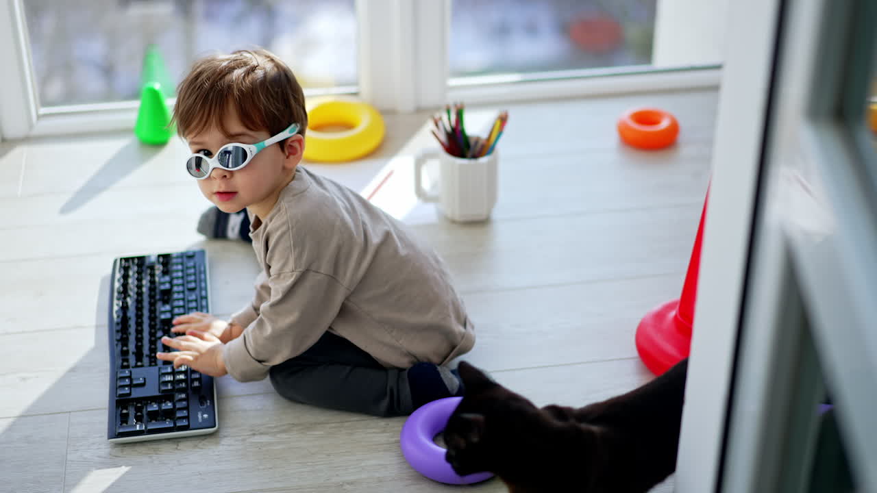 Baby boy playing with computer keyboard sitting on the floor. Black cat stands beside the child.
