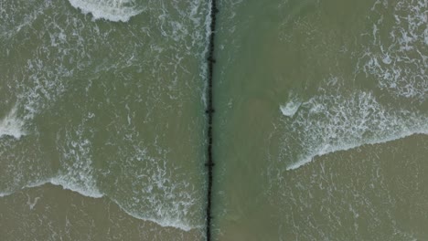 aerial birdseye view of an old wooden pier at the baltic sea coastline, overcast winter day, white sand beach, wooden poles, waves hitting shore, wide drone shot moving forward