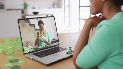 Smiling-african-american-woman-with-beer-wearing-clover-shape-band-on-video-call-on-laptop