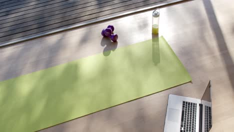 High-angle-view-of-yoga-mat,-laptop,-bottle-with-juice-and-weights-on-floor-at-home,-slow-motion