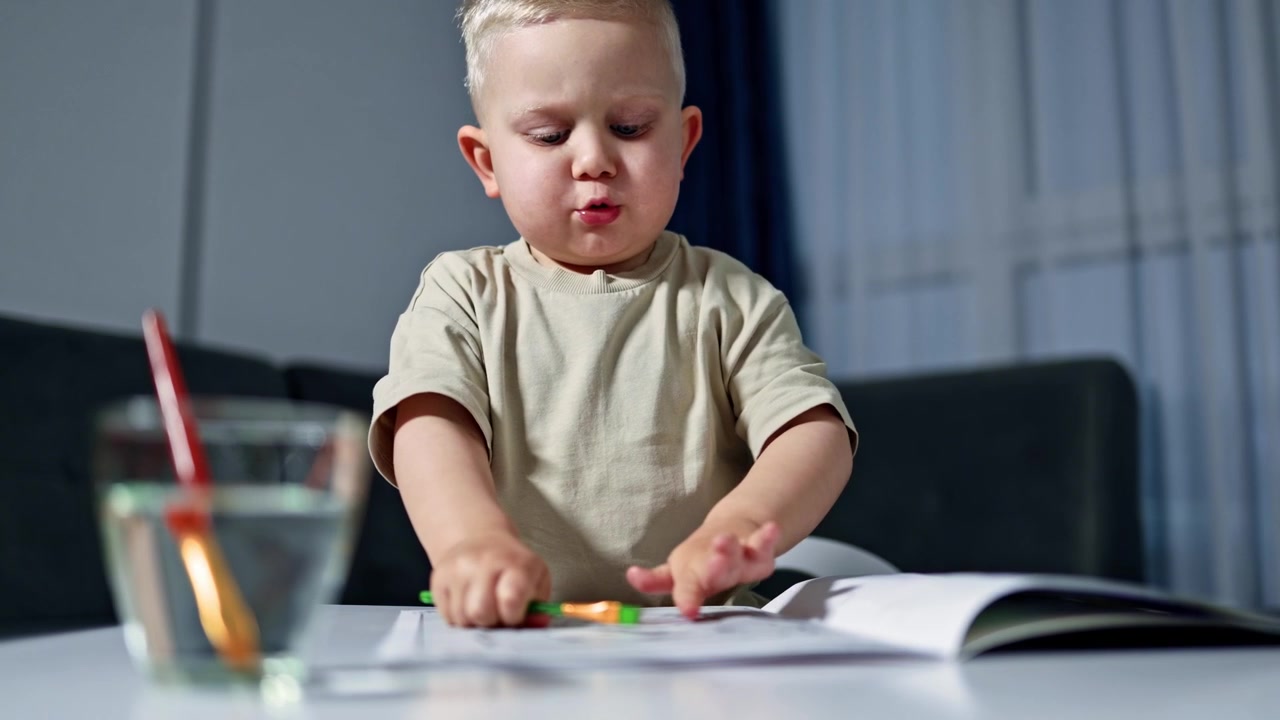 A 2 year old boy sits at a table and draws with a brush. The child is engaged in art and learns to draw with paints. Toddler learns to sit and draw with water.