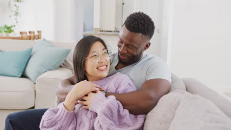 Diverse-couple-sitting-on-couch-and-embracing-in-living-room