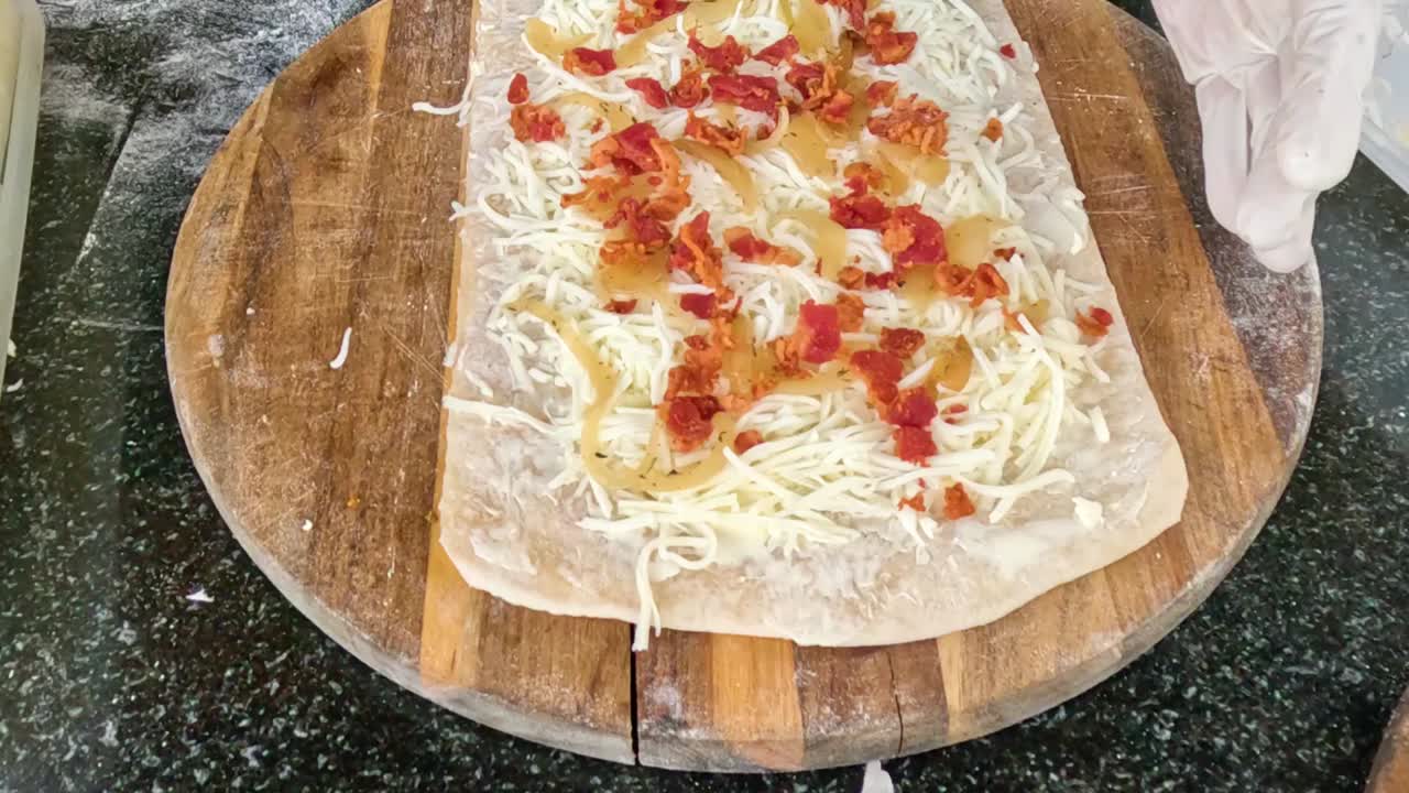 Gloved hands adding cheese and diced peppers to flatbread on a wooden board.
