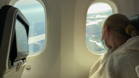 a child in a protective mask on his face looks out the window of the aircraft. traveling during the covid 19 epidemic