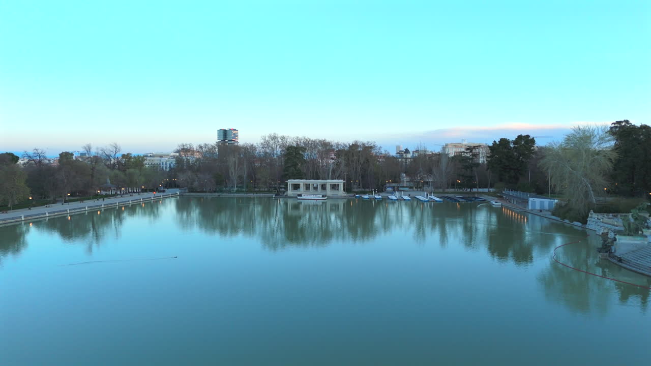 Stunning drone footage flying low over serene lake in Retiro Park during peaceful sunrise, capturing reflections, trees, boats, quiet pathways, and the tranquil atmosphere of nature and cityscape