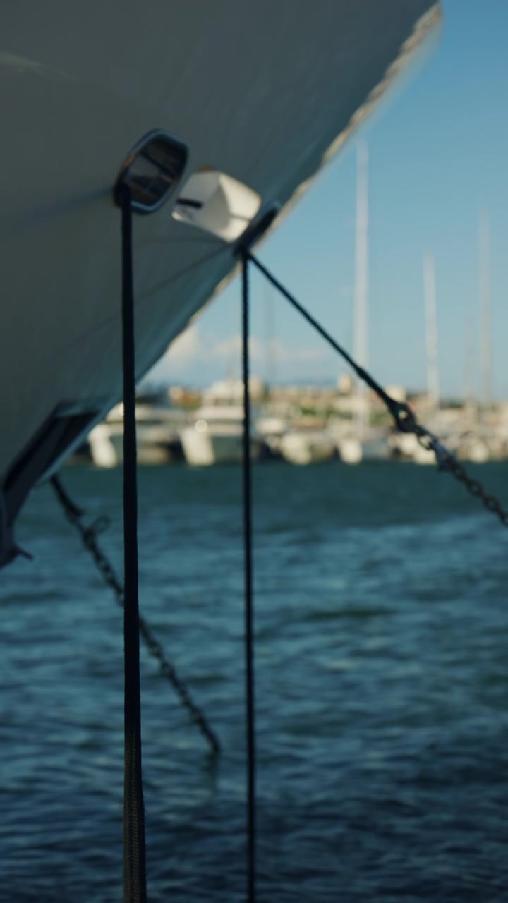 Nahaufnahme eines großen Yachtbugs mit Festmacherleinen und Ketten, mit Segelbooten und Marinagebäuden unscharf im Hintergrund.