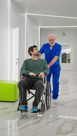 Vertical-video-of-a-confident-elderly-man-in-glasses-with-a-gray-beard-and-in-a-blue-doctors-uniform-carries-a-brunette-man-in-a-special-wheelchair-for-the-disabled-after-during-rehabilitation-after-fractures-in-a-modern-clinic