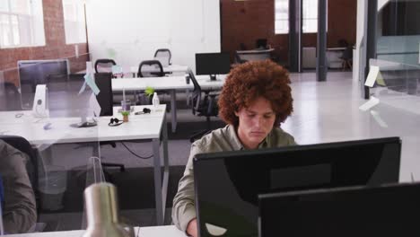 Mixed-race-businessman-sitting-at-desk-with-sneeze-guard-using-desktop-computer-looking-at-screen