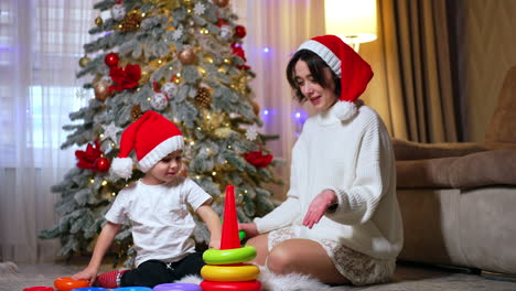 mother and child in red santa caps sit on the floor near the beautiful christmas tree. cute little boy plays with toy pyramid.
