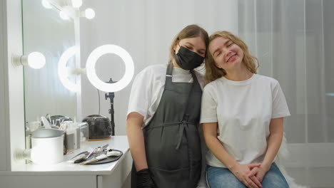 client and technician smile warmly at camera while leaning heads together inside modern beauty salon, with tools arranged on table beside mirror and soft white lighting enhancing cozy atmosphere