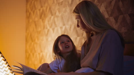 Mommy-strokes-hair-of-lovely-daughter-reading-book-on-bed