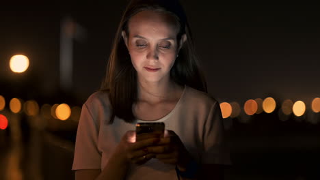 Una-Hermosa-Joven-En-Verano-En-Una-Gran-Ciudad-Nocturna-Mantiene-Un-Teléfono-Inteligente-En-Su-Interior-Y-Escribe-Un-Mensaje-Lee-Correos-Electrónicos-Y-Se-Comunica-En-Salas-De-Chat-Y-Redes-Sociales-Mantiene-Un-Blog-Mira-La-Pantalla-Del-Teléfono