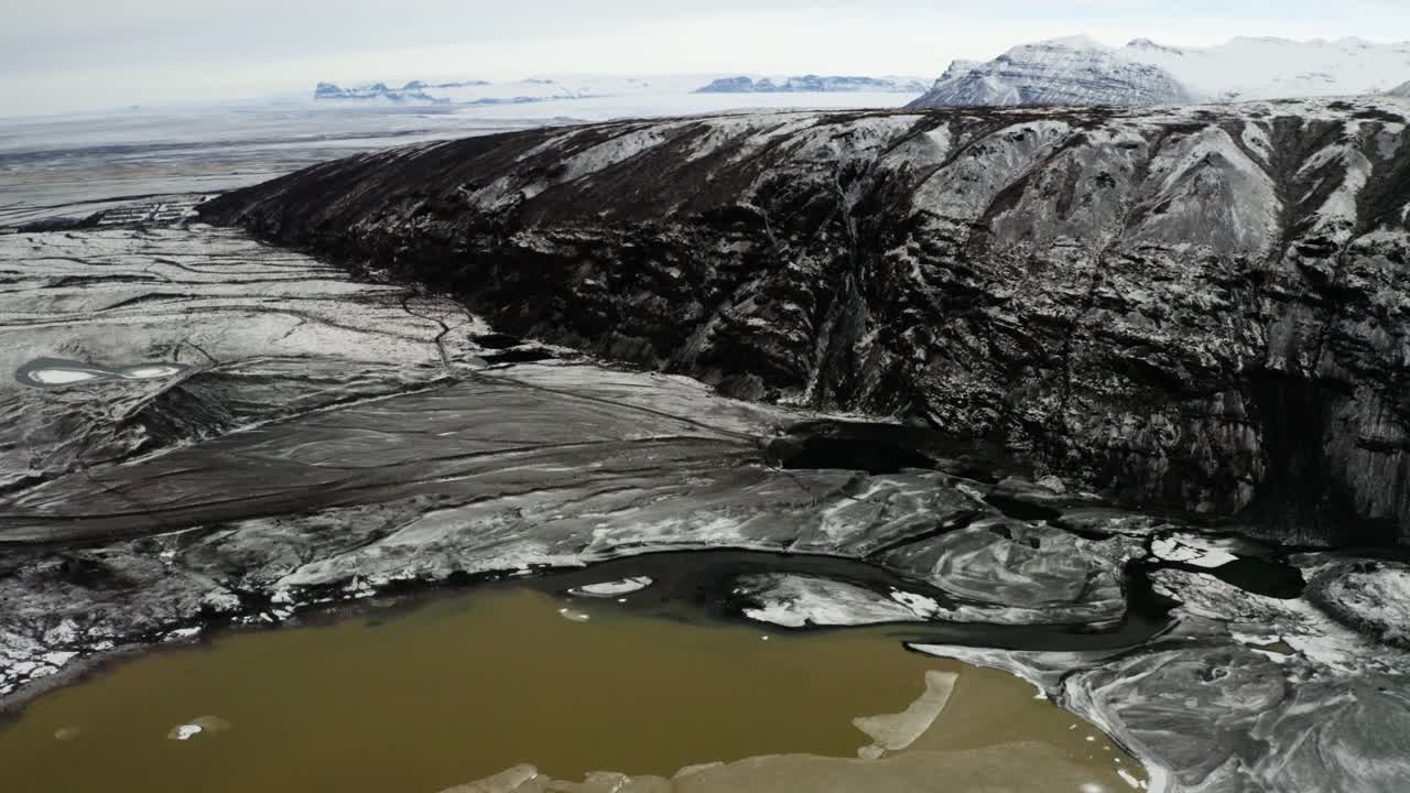 Captivating drone footage of the rocky cliffside and muddy meltwater lagoon across from Svinafellsjokull in Iceland.