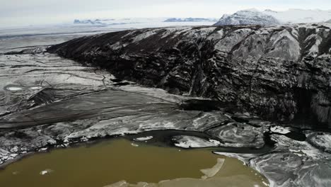 captivating drone footage of the rocky cliffside and muddy meltwater lagoon across from svinafellsjokull in iceland.
