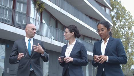 smiling confident business colleagues walking at office building, drinking coffee and talking together