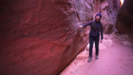 Wide-shot-of-a-female-hiker-walking-backwards-rubbing-her-fingers-along-the-sandstone-wall-of-a-slot-canyon-while-gazing-upward,-Slow-Motion-shot