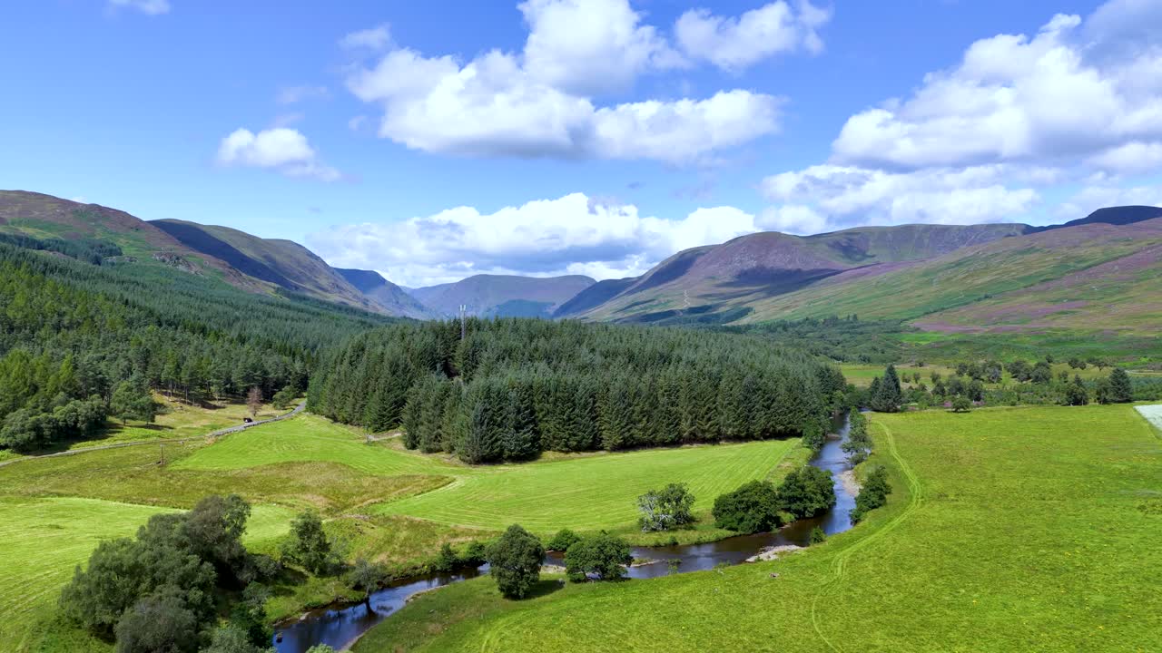 Drone glides above green river valley, forest, and mountains under bright daylight and blue sky
