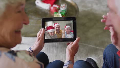 Sonriente-Pareja-De-Ancianos-Caucásicos-Usando-Una-Tableta-Para-Una-Videollamada-Navideña-Con-La-Familia-En-La-Pantalla