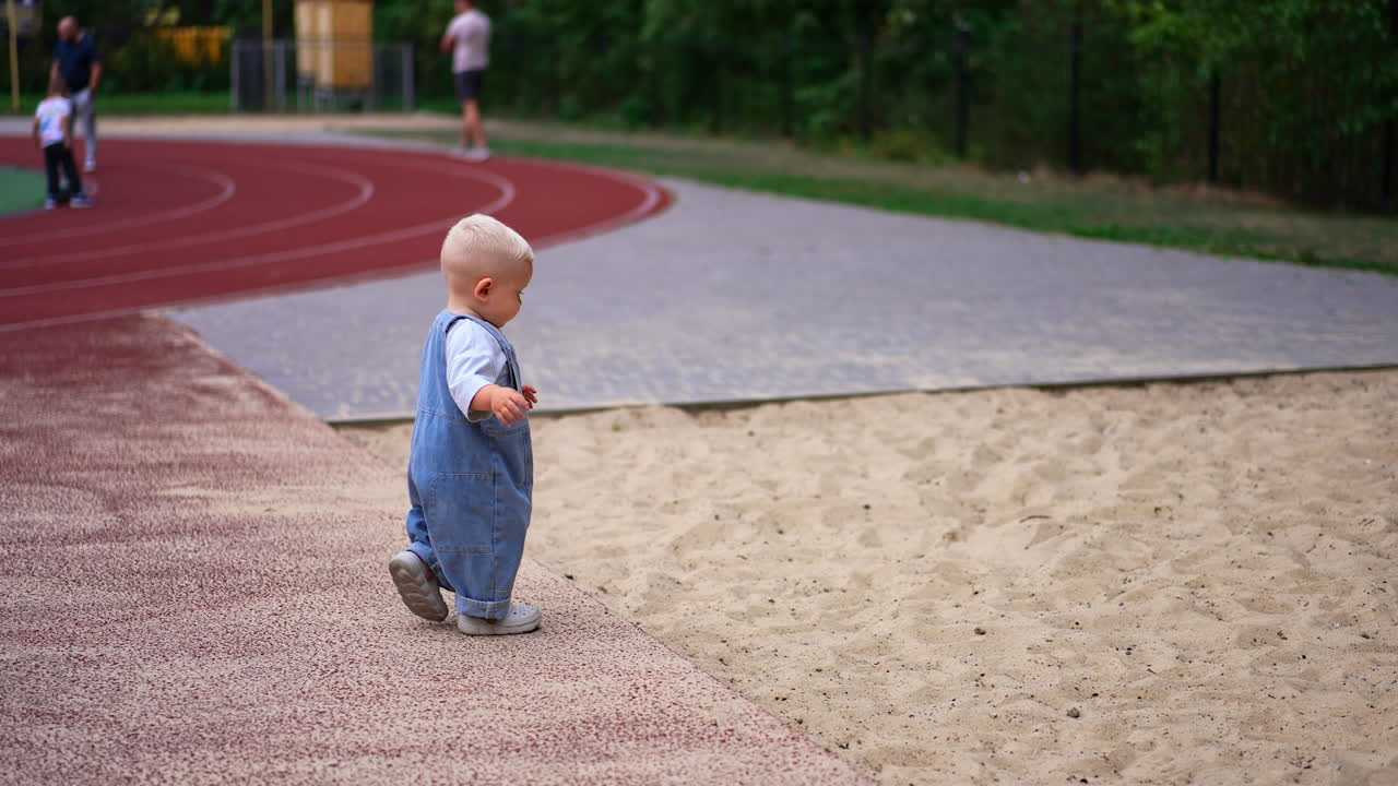 Cute little baby in loose jeans romper is outdoors. Kid steps into the sand and bends to pick some.