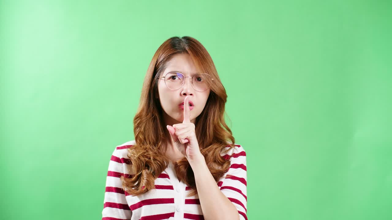 안경을 쓴 젊은 아시아 여성이 당신의 몸짓을 보고 눈을 가리키며 녹색 화면 배경을 주시합니다.