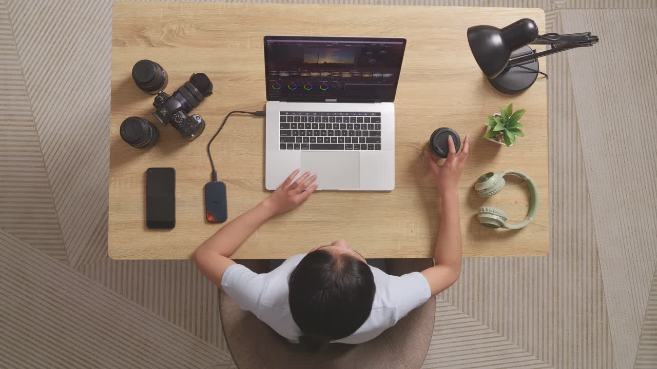 Top View Of Asian Woman Color Grading Drinking Coffee While Sitting In The Workspace Using A Laptop Next To The Camera Editing The Video At Home