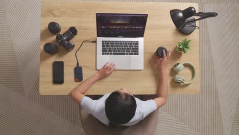 top view of asian woman color grading drinking coffee while sitting in the workspace using a laptop next to the camera editing the video at home