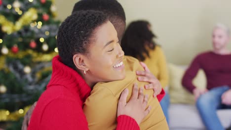 Happy-diverse-male-and-female-friend-hugging-in-front-of-christmas-tree-at-home