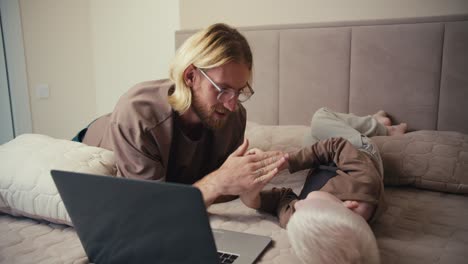 A-happy-blond-man-in-glasses-with-a-beard-plays-with-his-little-albino-son-with-white-hair-color-in-rock-paper-scissors-and-then-turns-on-a-cartoon-on-a-gray-laptop-while-spending-time-together-in-the-room