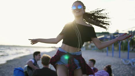 mujer joven hipster despreocupada con dreads bailando en la puesta de sol con un grupo de amigos sentados en sillas fáciles en la playa y tocando la guitarra en una noche de verano durante una puesta de sol. disparo en cámara lenta