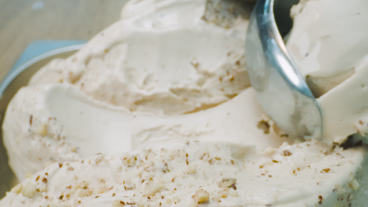 A close-up of a creamy ice cream with hazelnut chunks being scooped out using a scooper. The rich texture and visual contrast between the smooth ice cream and the crunchy hazelnut.