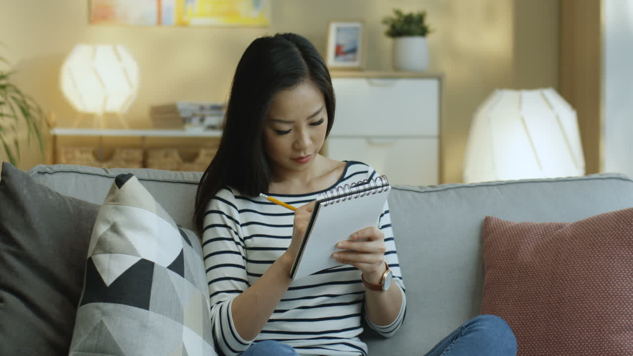 Young Woman In Striped Shirt Taking Notes On Notebook And Thinking Sitting On The Sofa In The Living Room