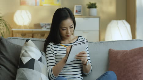Young-Woman-In-Striped-Shirt-Taking-Notes-On-Notebook-And-Thinking-Sitting-On-The-Sofa-In-The-Living-Room