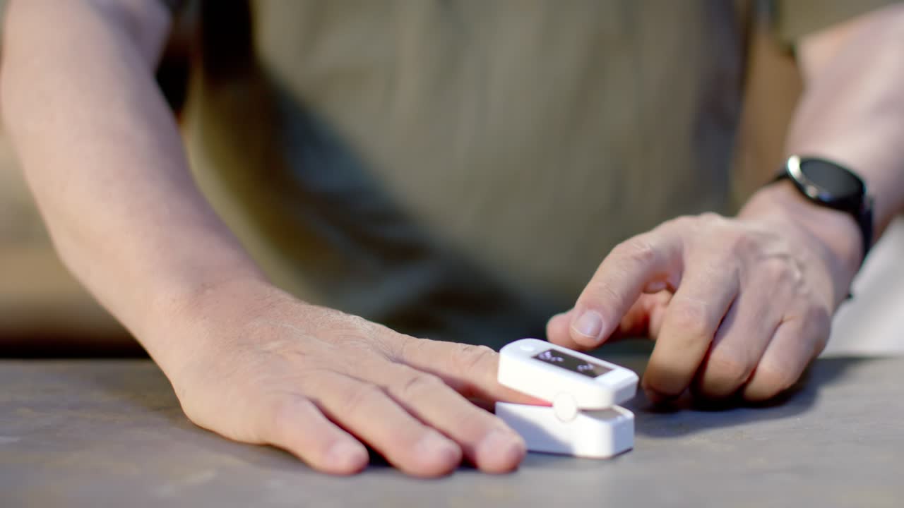 Senior Asian male bringing white oximeter to left finger, clipping and checking oxygen on table