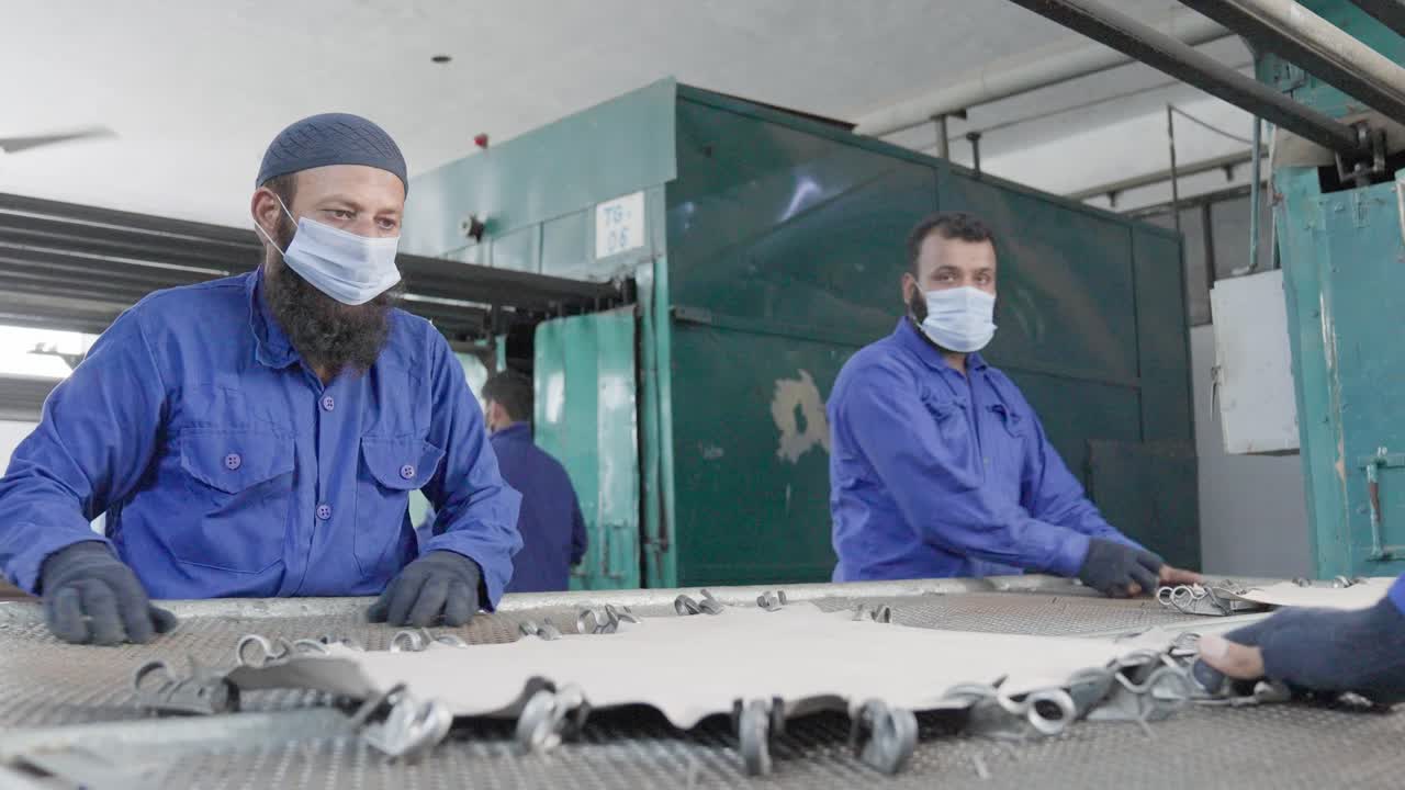 Worker in blue clothes and masks of a leather factory working with the animal skin, attach with brackets on metal, turn around, static shot